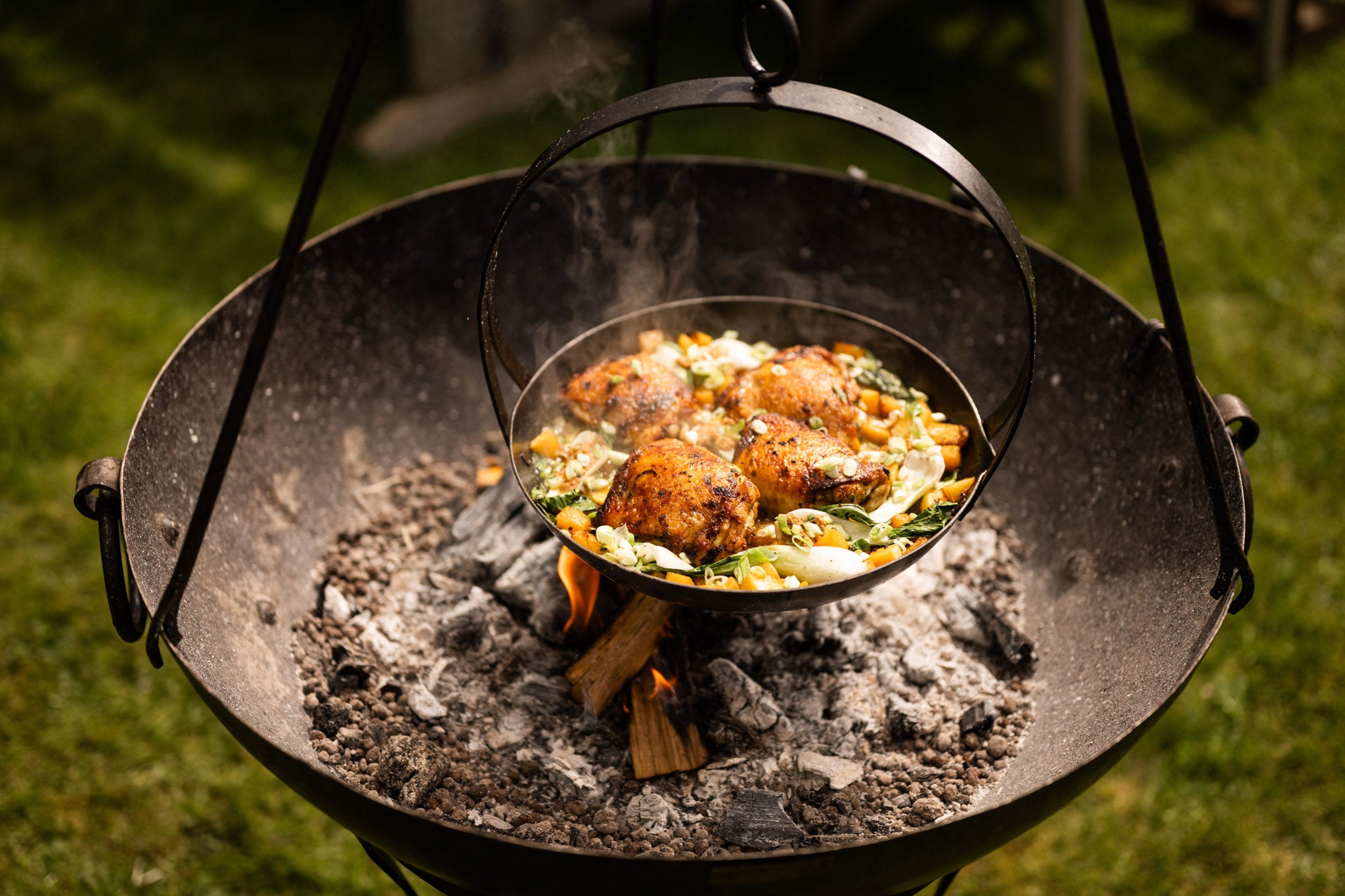 Tripod skillet suspended over an open fire, cooking seasoned chicken and vegetables in an outdoor fire pit.