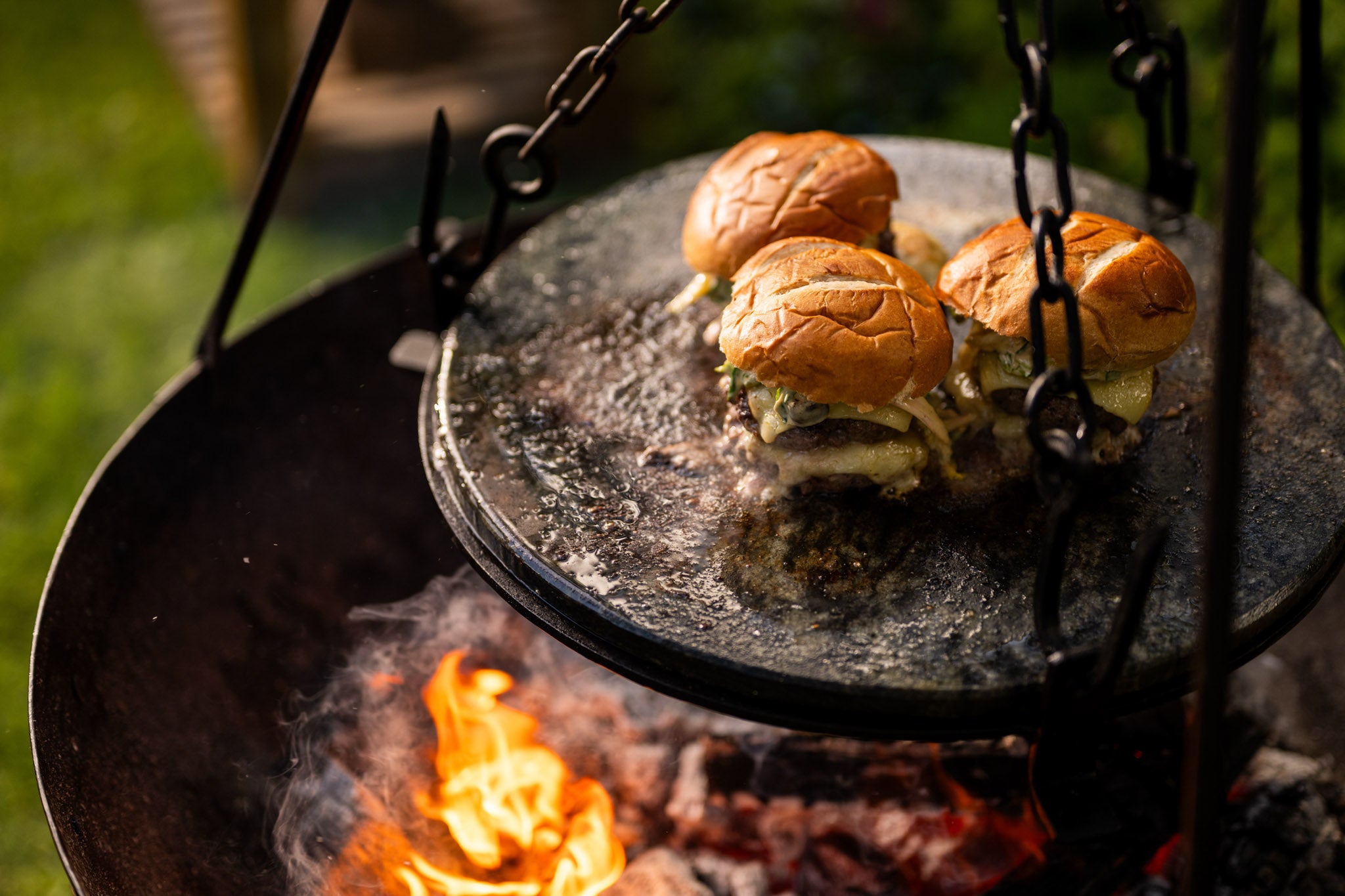 Cheeseburgers cooking on a hot stone griddle plate suspended above glowing coals and open flames in an outdoor fire pit.