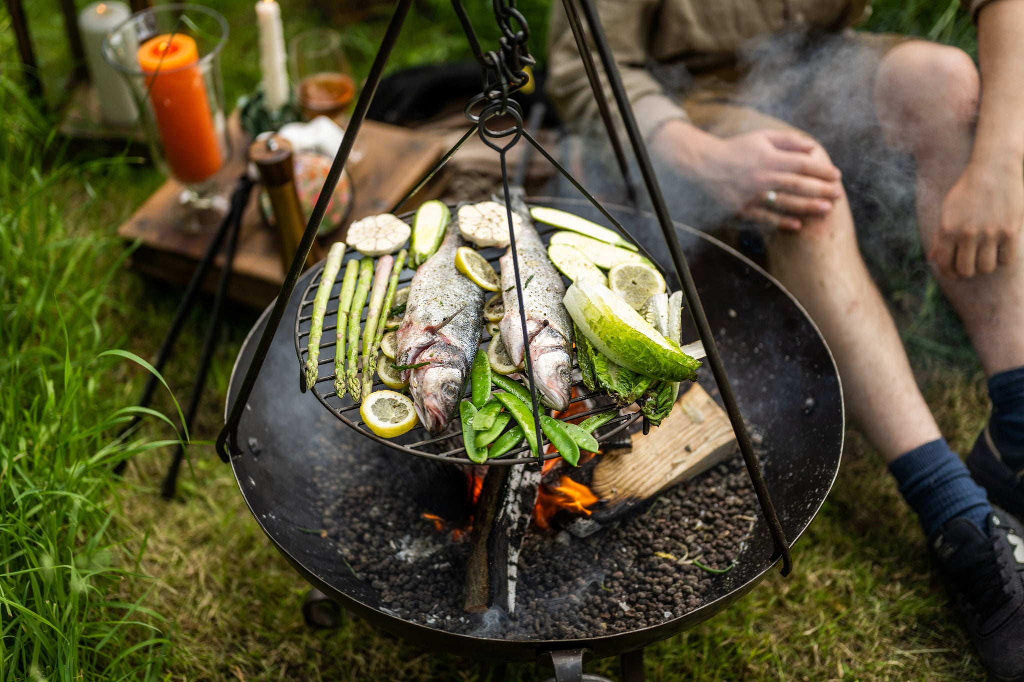 Whole fish and fresh vegetables grilling over an open fire on a suspended BBQ cooking tripod in an outdoor campsite setting.