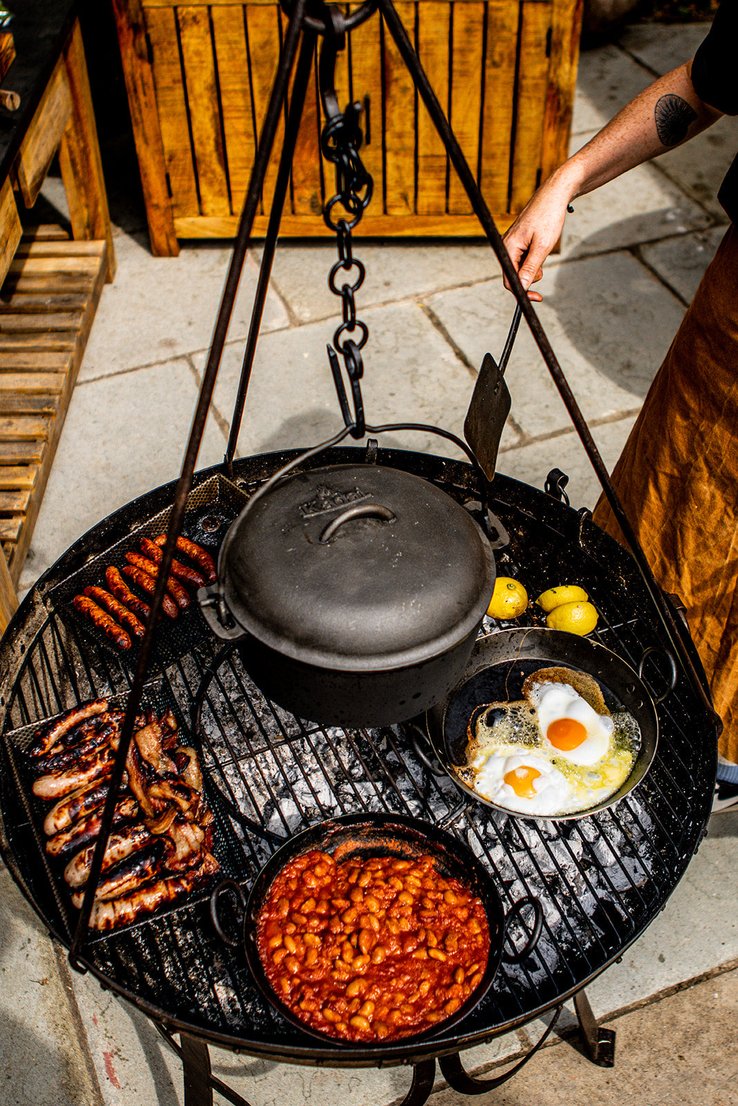 A round charcoal grill setup outdoors with a hanging cast iron dutch oven, sausages, eggs, and a bowl of baked beans, with a person's arm visible adjusting the hanging mechanism.