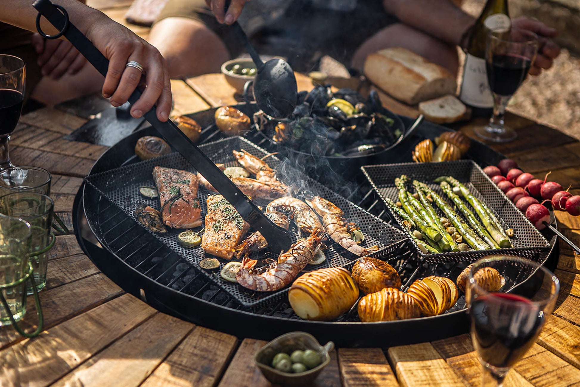 Outdoor cooking scene with grill, food, and people around a table.