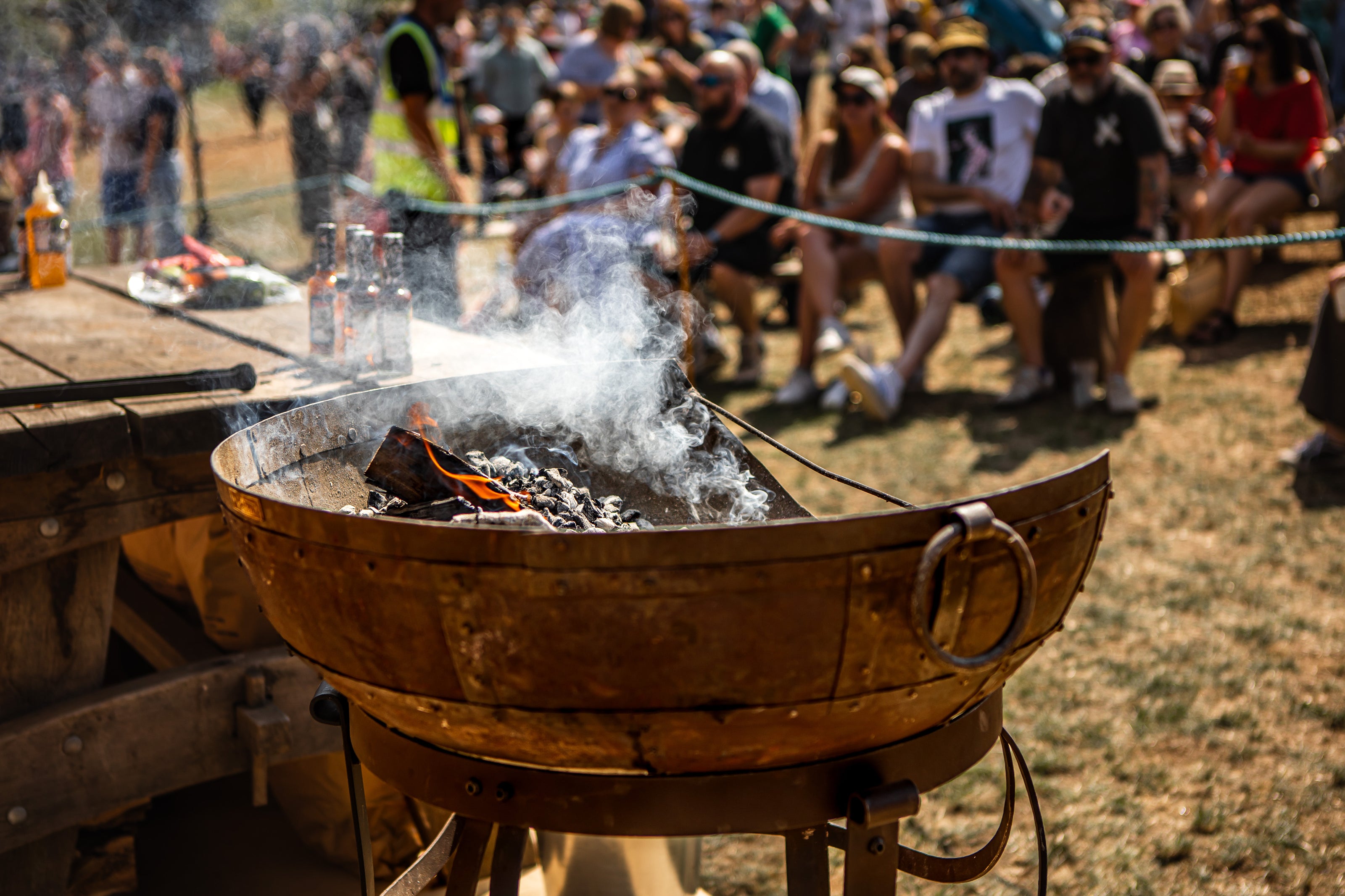 Kadai fire pit with flames and smoke at a outdoor event with people in the background.