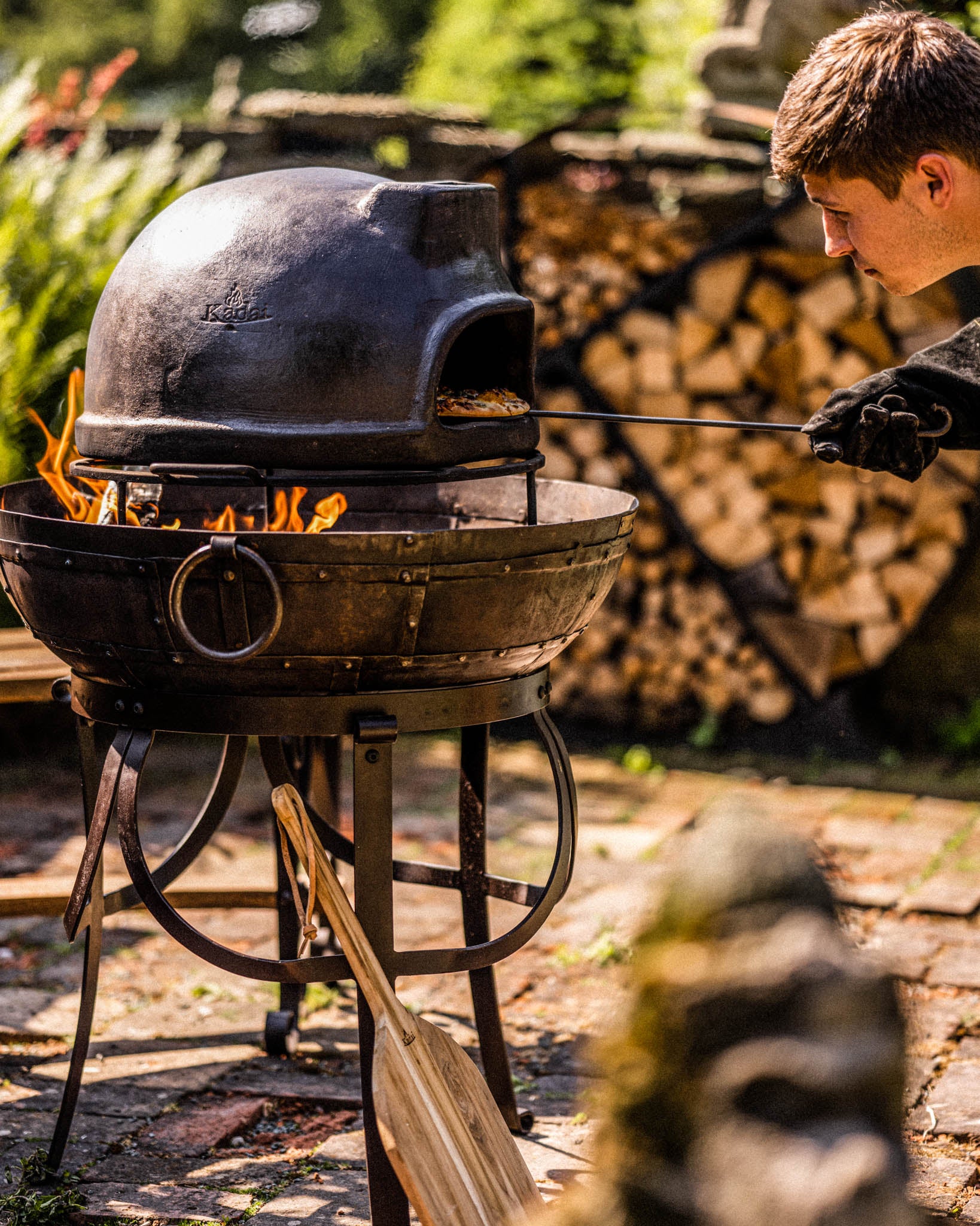 Outdoor wood fired oven in use, with a person checking pizza over a Kadai fire pit in a garden setting