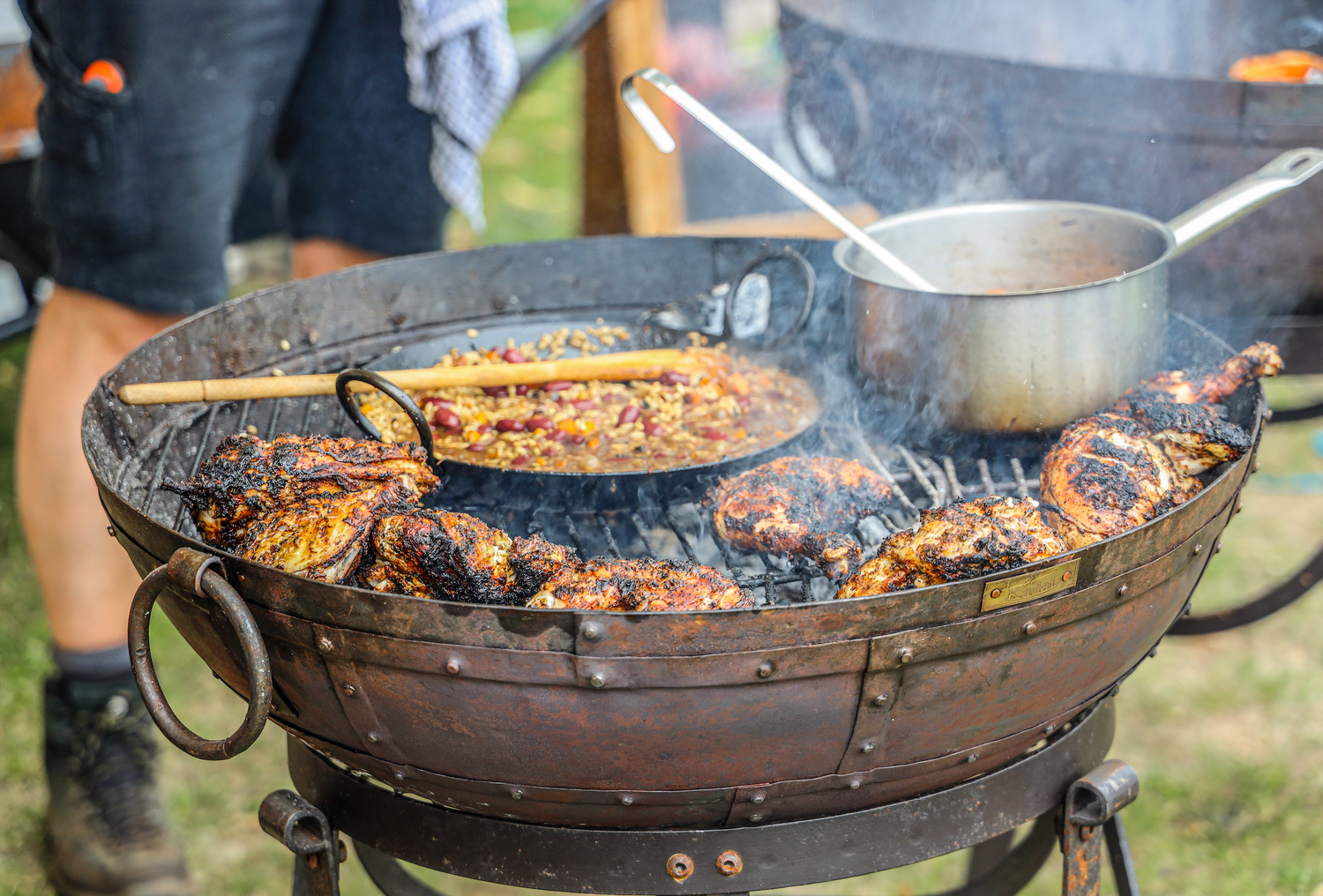 Outdoor cooking scene with a large metal Kadai grill and food being cooked.