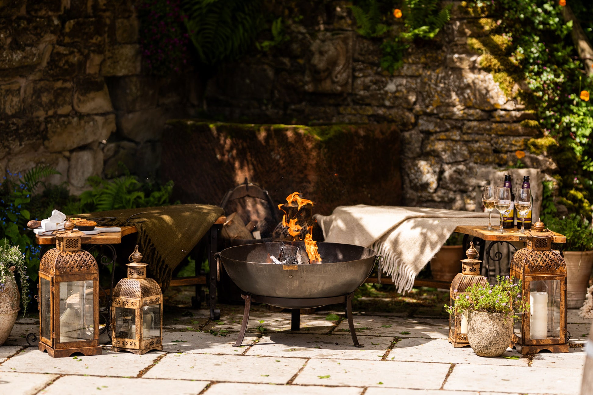 Outdoor setting with a kadai contemporary fire pit, lanterns, and decorative items against a stone wall.
