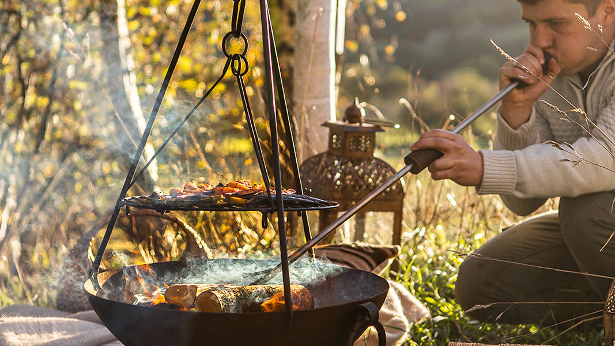 Person cooking over an open fire using bloker to increase flames and heat in a natural setting.