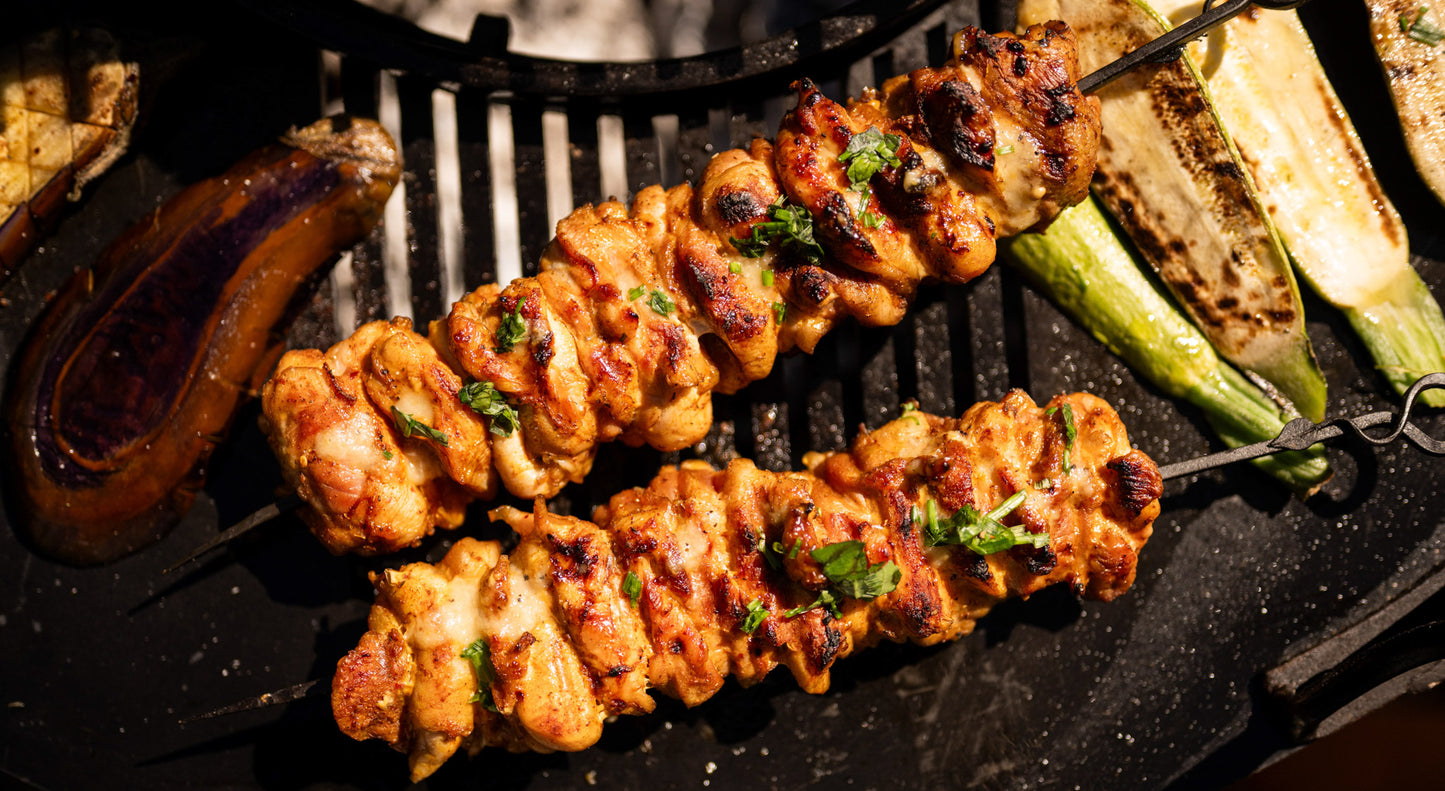 Close-up of grilled chicken skewers and roasted vegetables on a Plancha Kadai Set over charcoal.