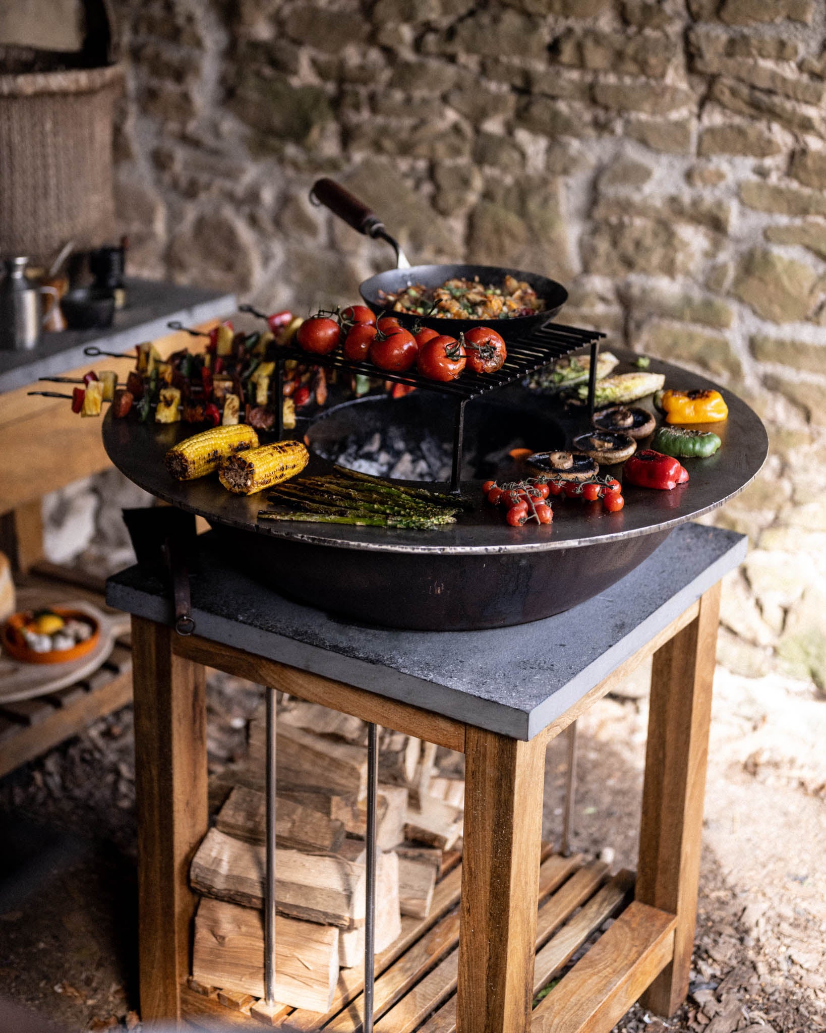 Round BBQ plancha grill on a wooden outdoor stand, cooking assorted vegetables including corn, asparagus, tomatoes, mushrooms, peppers, and skewers around an open fire center.