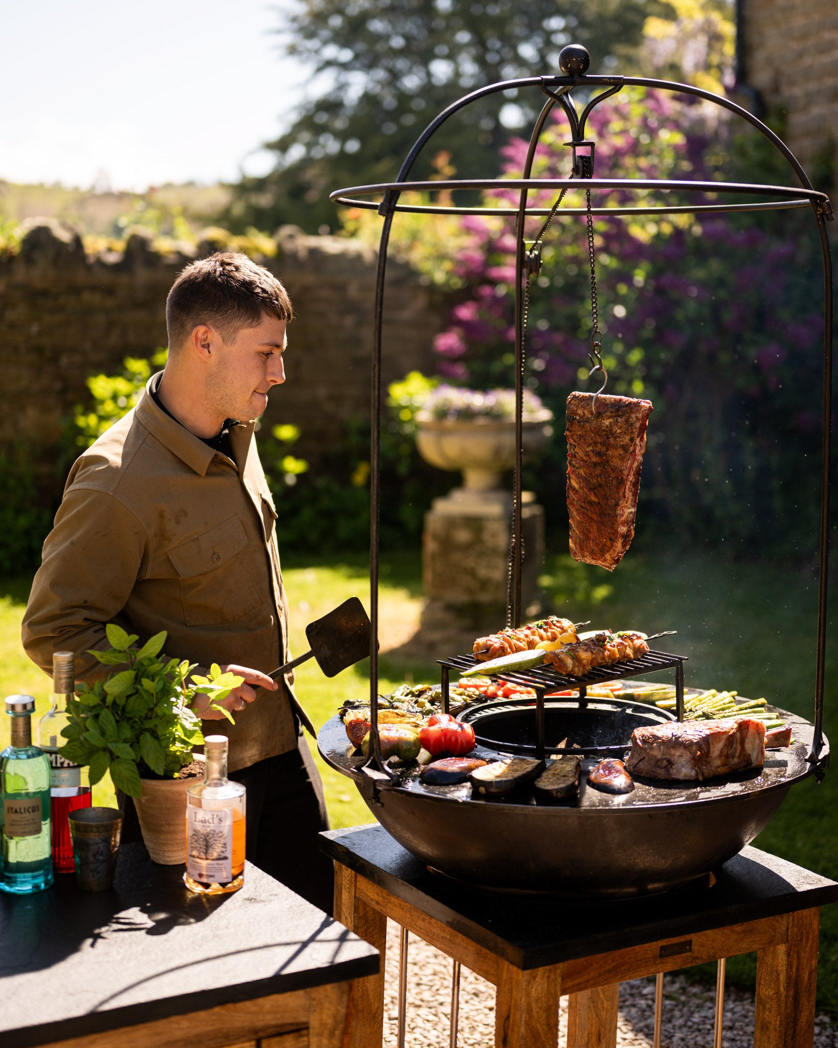 Plancha Kadai Set grill with various meats and vegetables cooking on it, including a hanging piece of meat, surrounded by greenery and garden furniture.