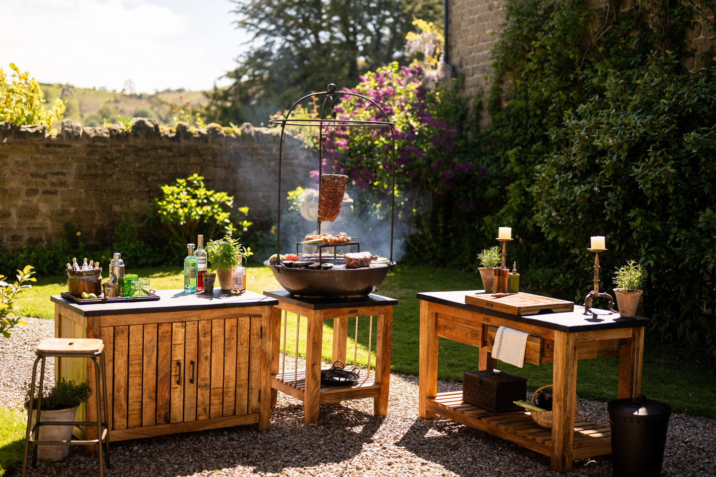 Outdoor Plancha Kadai Set with hanging meat, grilled vegetables, and skewers, surrounded by wooden tables and garden plants.