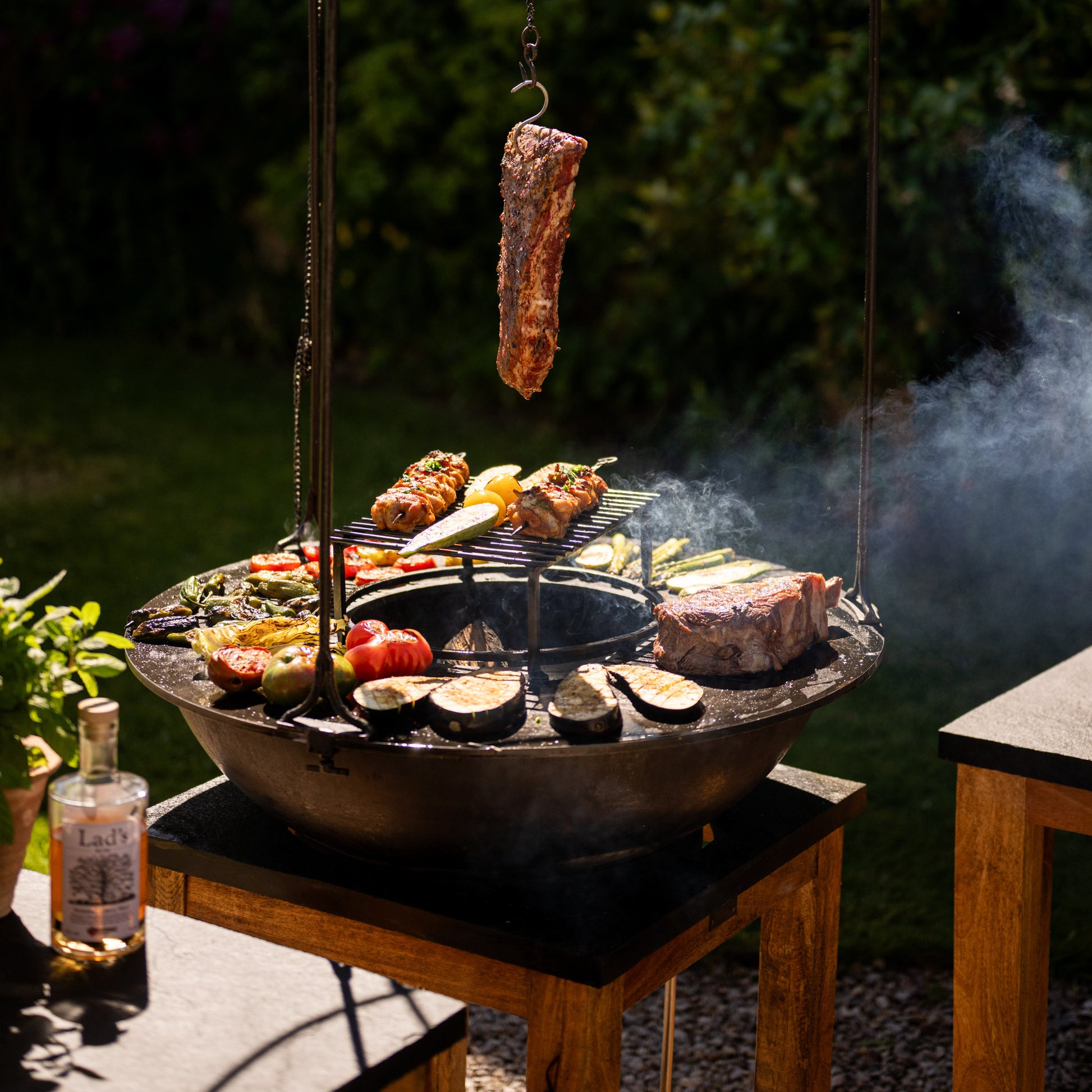 Plancha Kadai Set grilling an assortment of meats and vegetables outdoors, with a hanging rib, skewers, and charred vegetables on a sunny garden setup.
