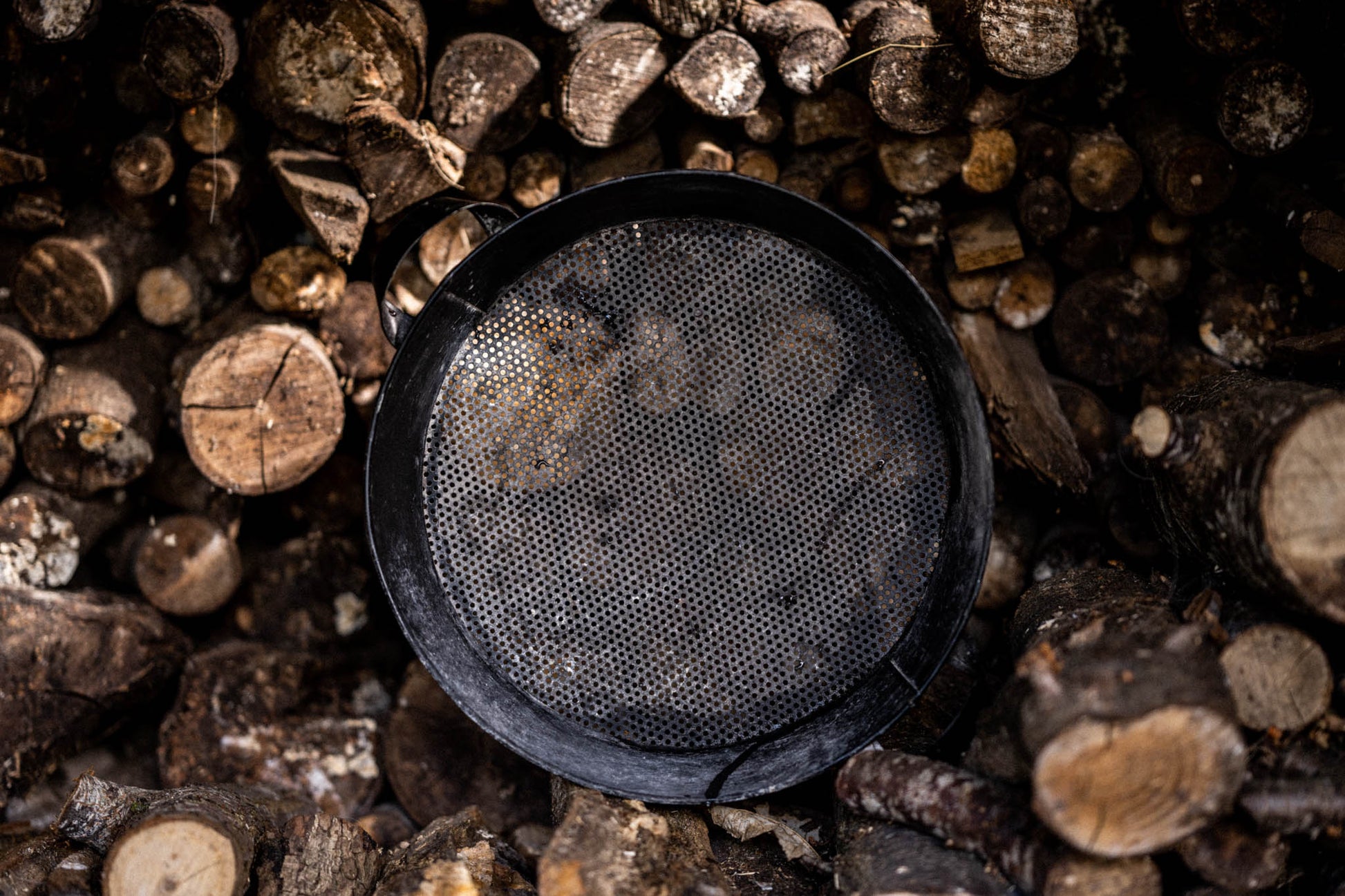 Top-down view of a black steel Kadai riddle sieve placed on a log pile, used for cleaning fire pit beads and embers