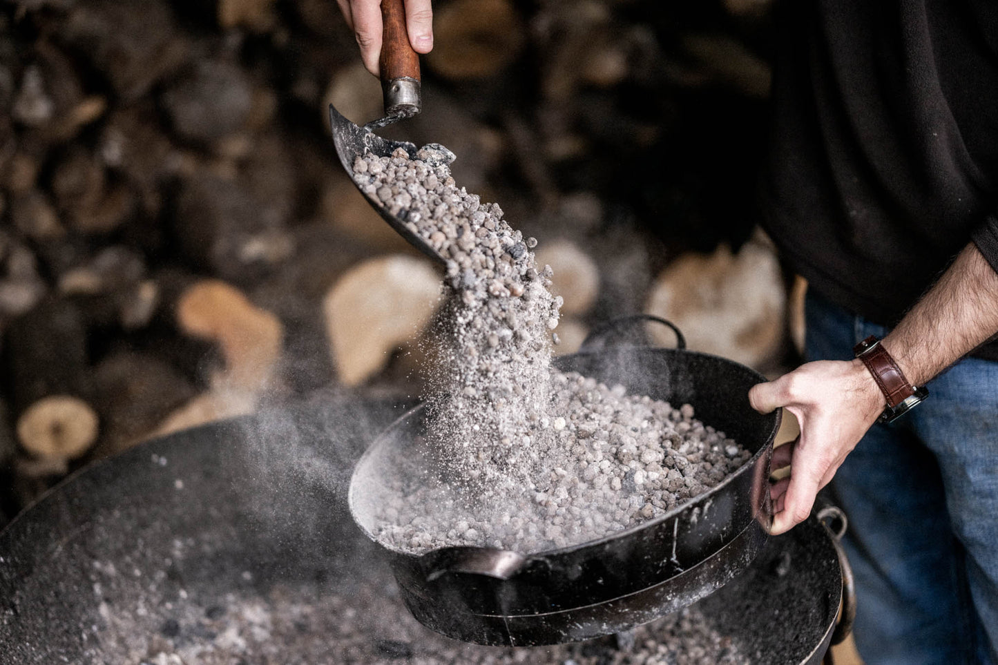 Person pouring hot ash and embers from a Kadai shovel into a black metal ash pan, with dust and smoke rising in the air.