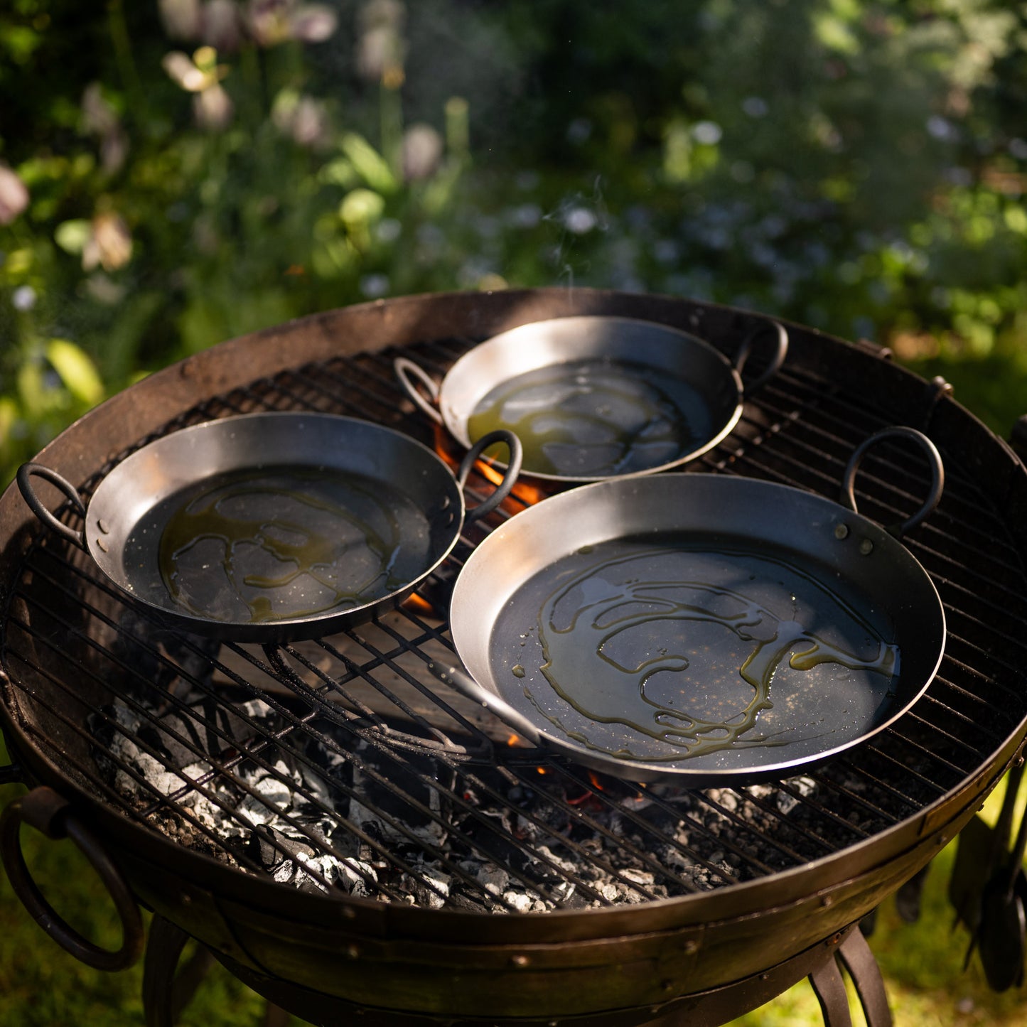 Three BBQ skillets heating with olive oil on an outdoor grill above glowing charcoal and open flames.