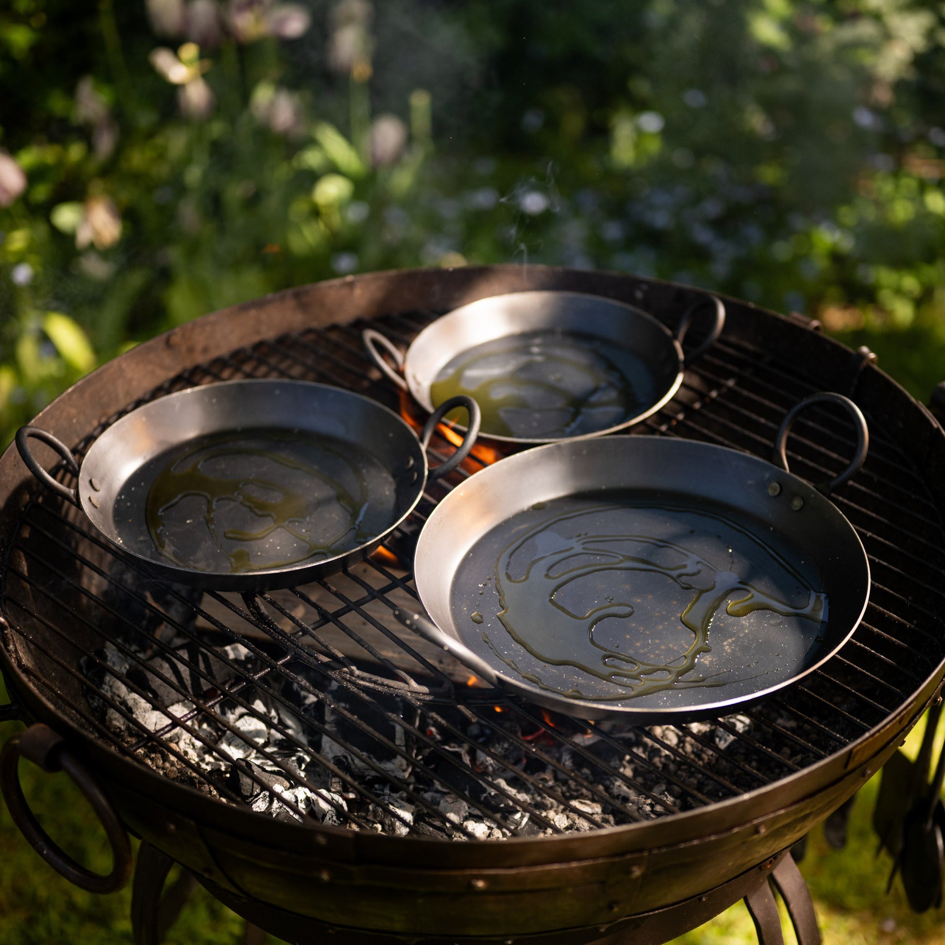 Three BBQ skillets heating with olive oil on an outdoor grill above glowing charcoal and open flames.