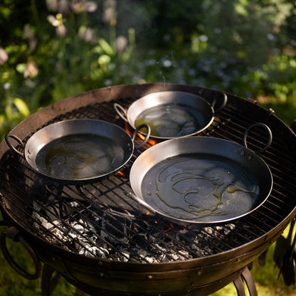 Three BBQ skillets heating with olive oil on an outdoor grill above glowing charcoal and open flames.