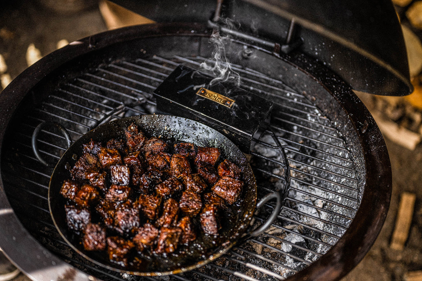 Smoker box on a grill with a cast iron pan of cooked meat, smoke rising.
