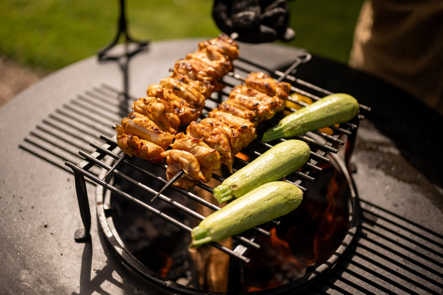 Chicken BBQ skewers and fresh zucchini grilling over open flames on a metal barbecue rack outdoors.