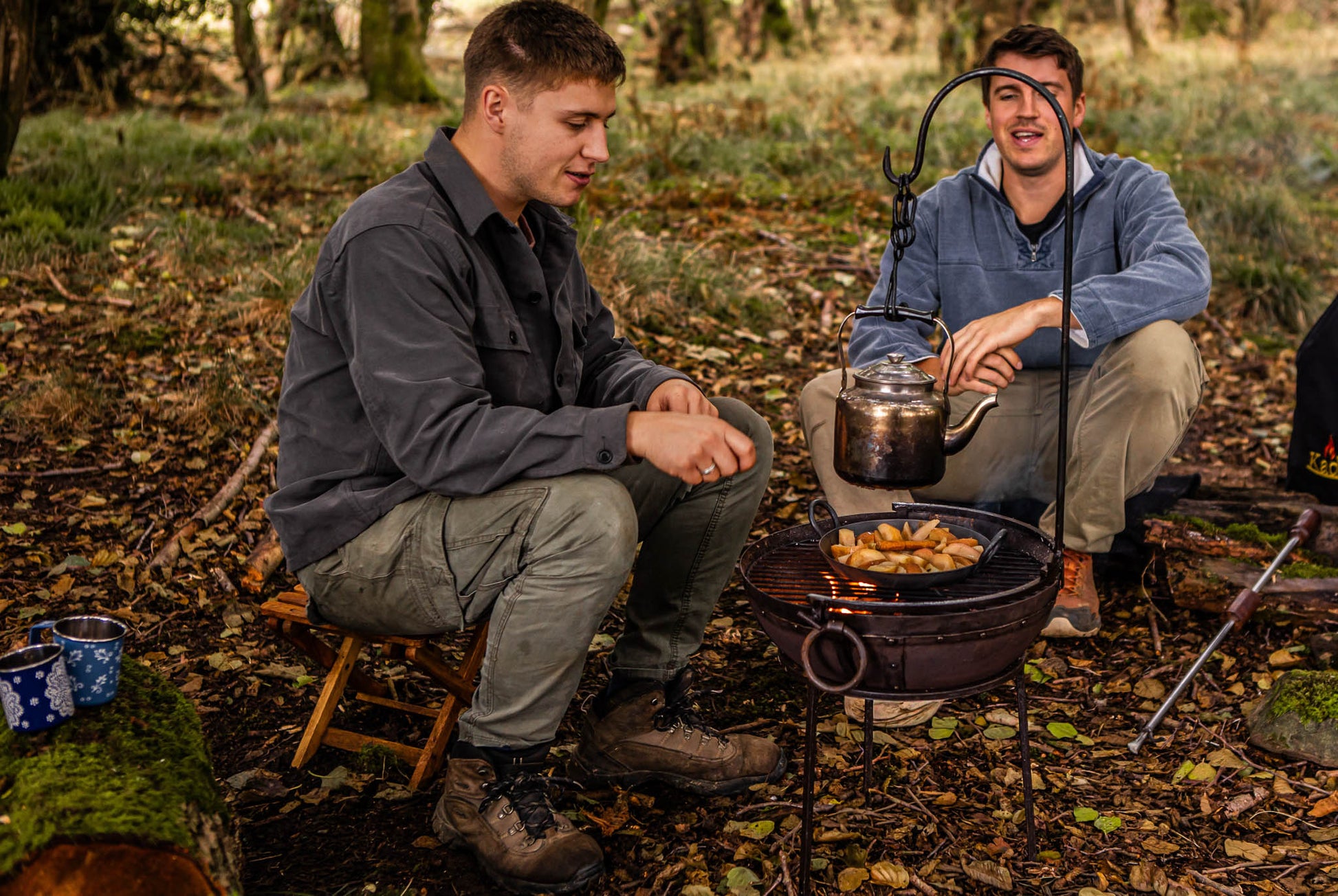 Portable BBQ hook holding a kettle over a fire pit, with two people cooking outdoors in a woodland setting