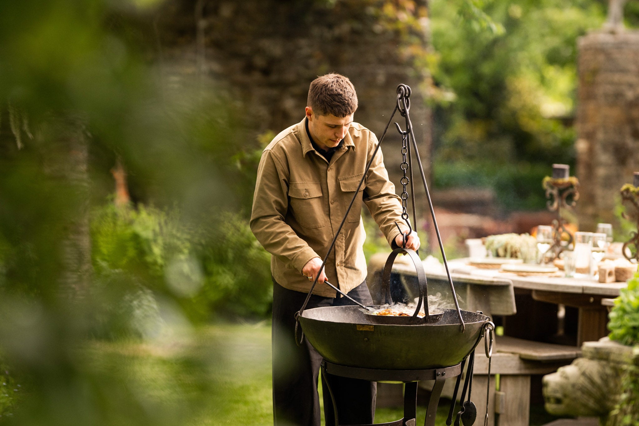 Person cooking outdoors with a hanging tripod skillet over an open fire pit in a garden dining setting.