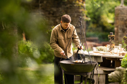 Person cooking outdoors with a hanging tripod skillet over an open fire pit in a garden dining setting.