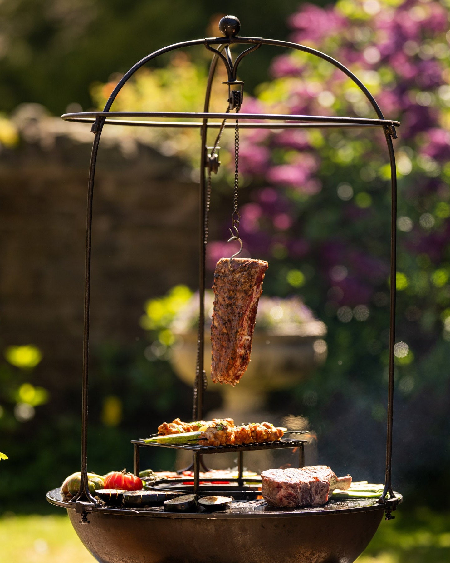 Outdoor BBQ plancha grill with a suspended rack of ribs hanging over live fire, surrounded by steaks, skewers, and grilled vegetables on a circular cooking surface in a sunlit garden