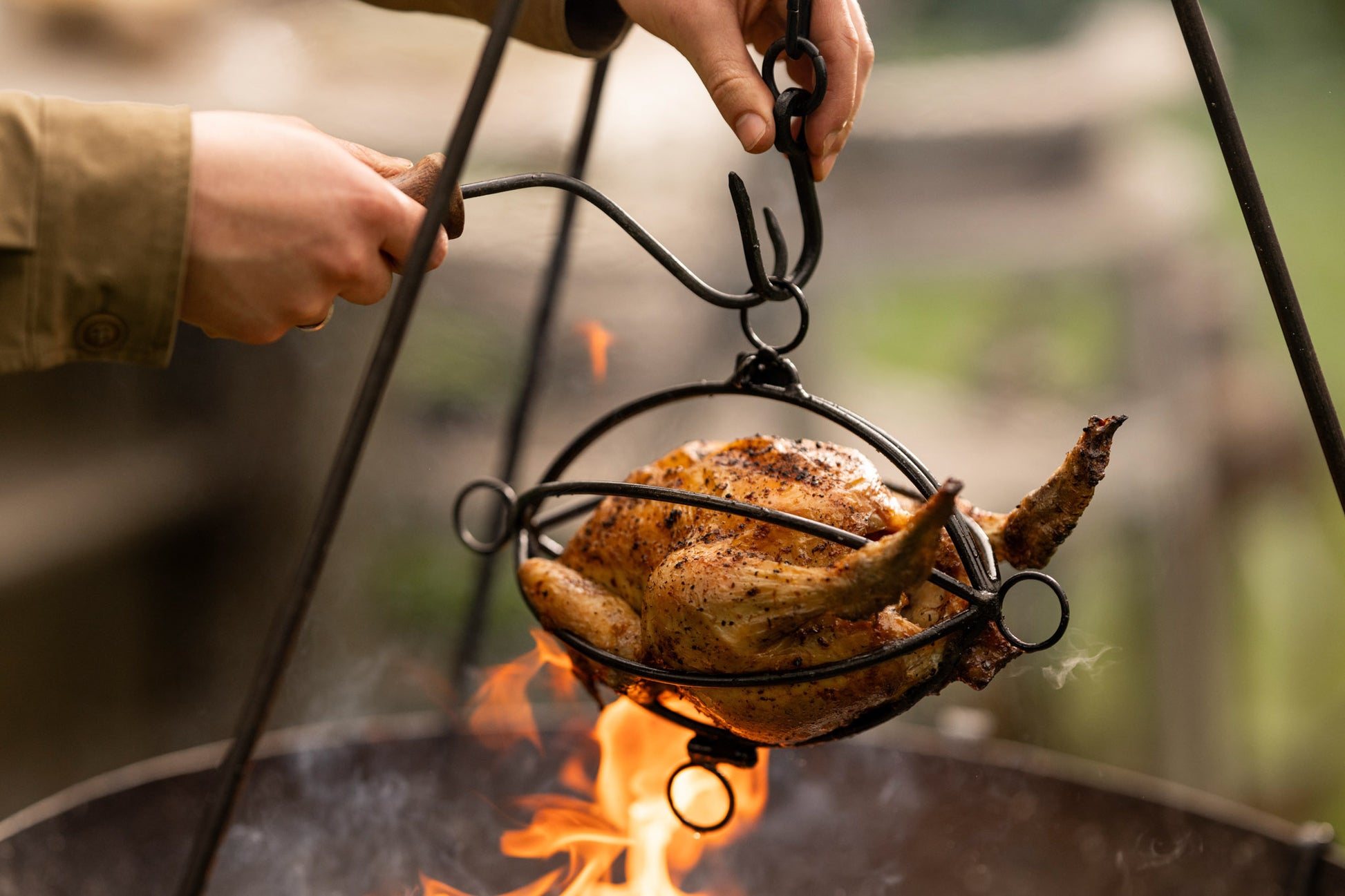 Whole roasted chicken suspended over an open fire in a tripod roaster basket during outdoor cooking.