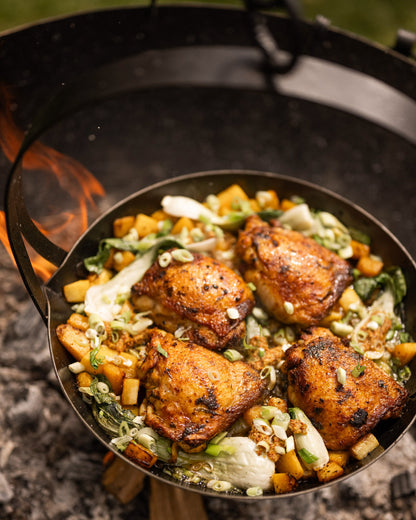 Tripod skillet suspended over a Kadai firebowl barbecue with roasted chicken and vegetables cooking above open flames outdoors