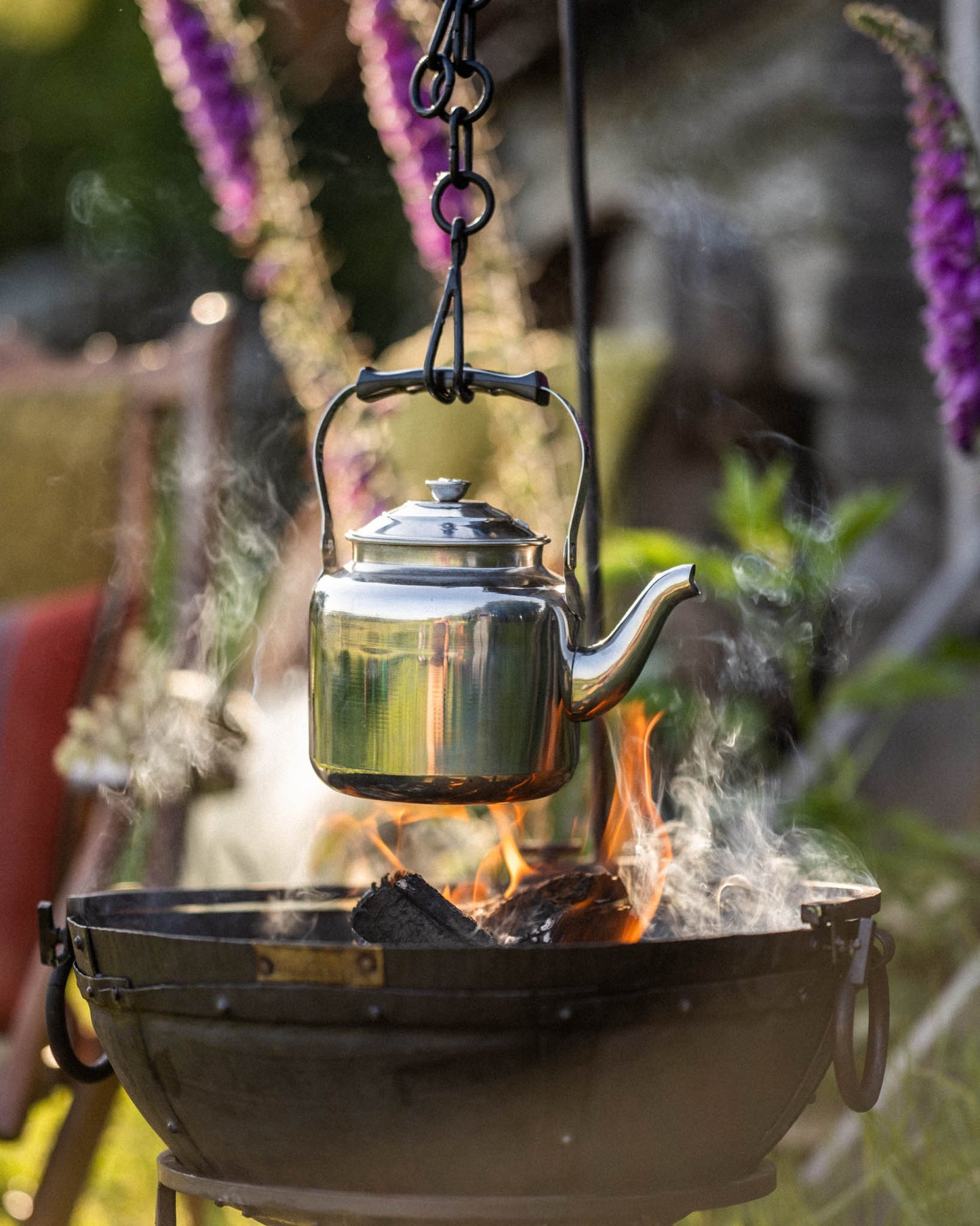 Camping BBQ kettle hanging over an open fire pit, with steam rising as it heats above glowing embers