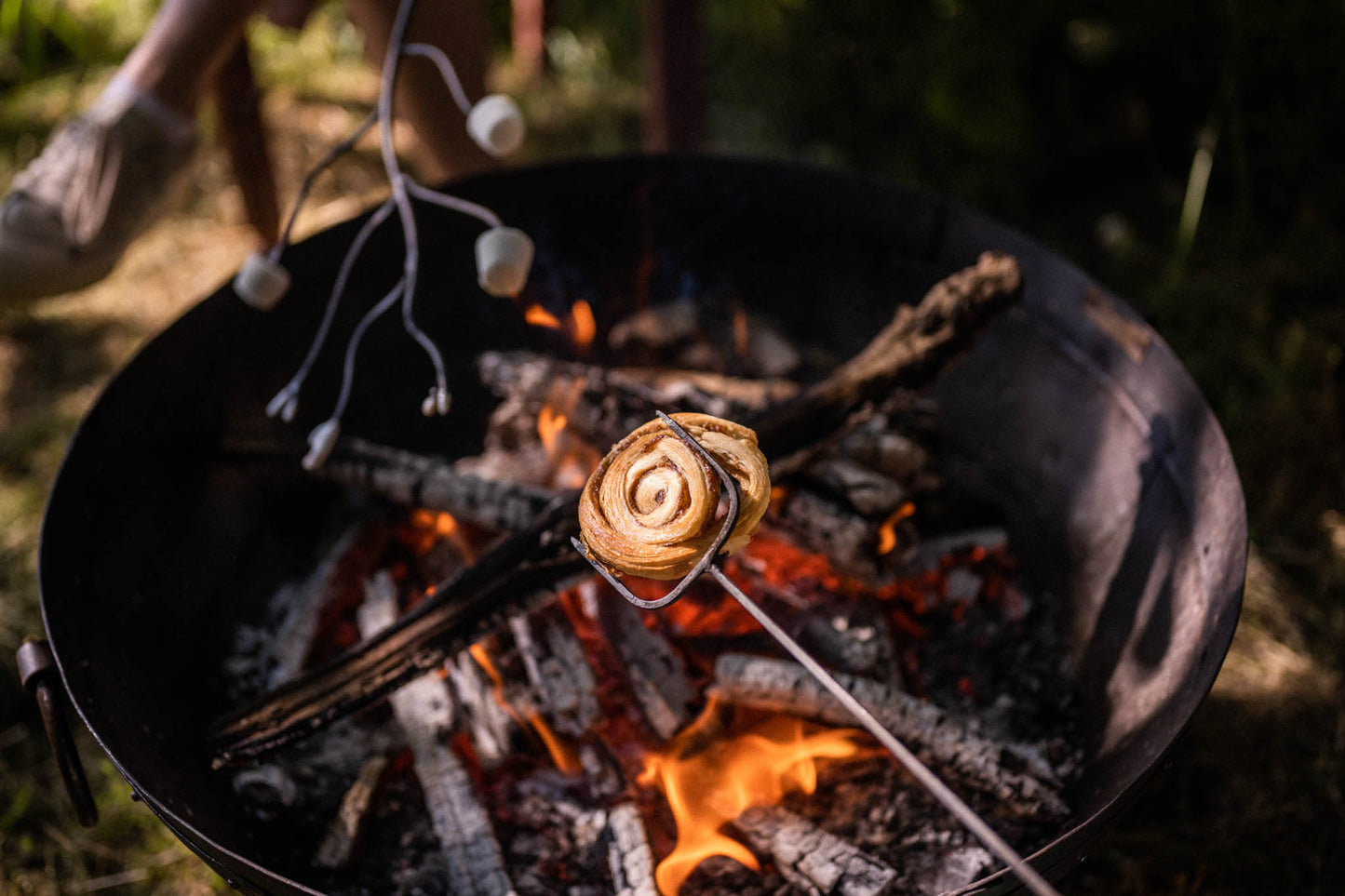 Marshmallow toasting fork holding a cinnamon pastry over glowing embers in a Kadai firebowl, with marshmallows roasting over an open outdoor fire at dusk