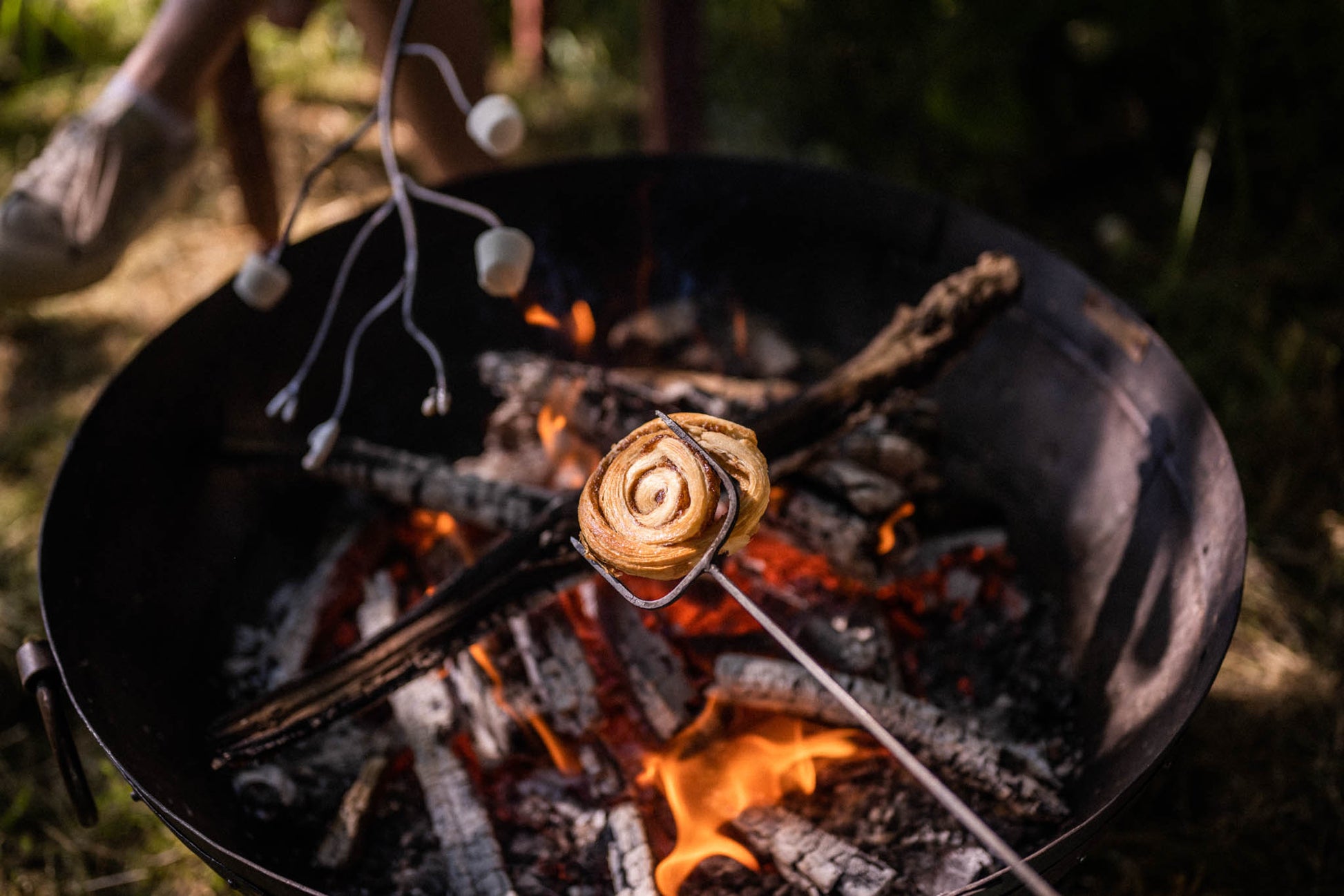 Marshmallow toasting fork holding a cinnamon pastry over glowing embers in a Kadai firebowl, with marshmallows roasting over an open outdoor fire at dusk
