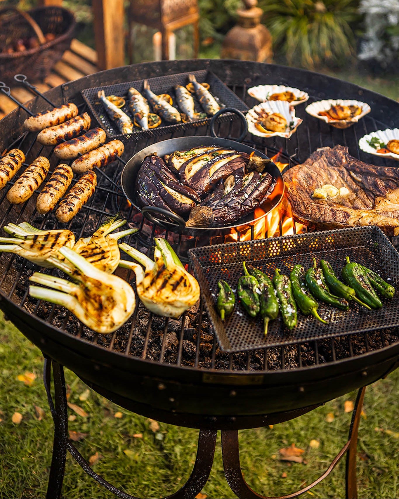 Medium sized BBQ fire pit with Kadai grill cooking steak, sausages, fish and vegetables over open flames