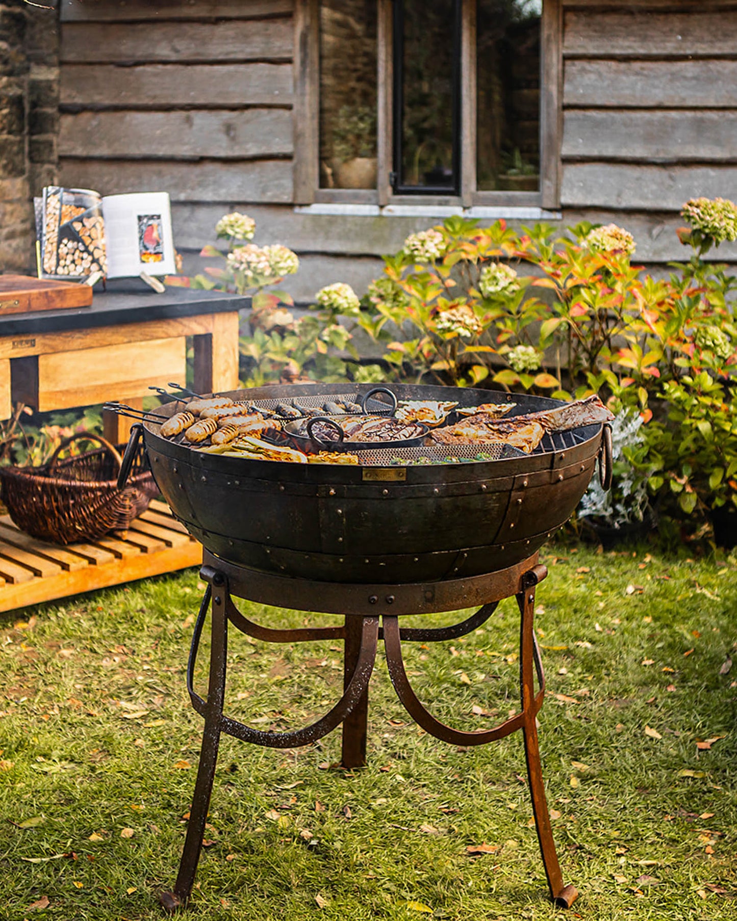 Medium sized BBQ fire pit with Kadai grill cooking sausages and meat in a garden beside a wooden house