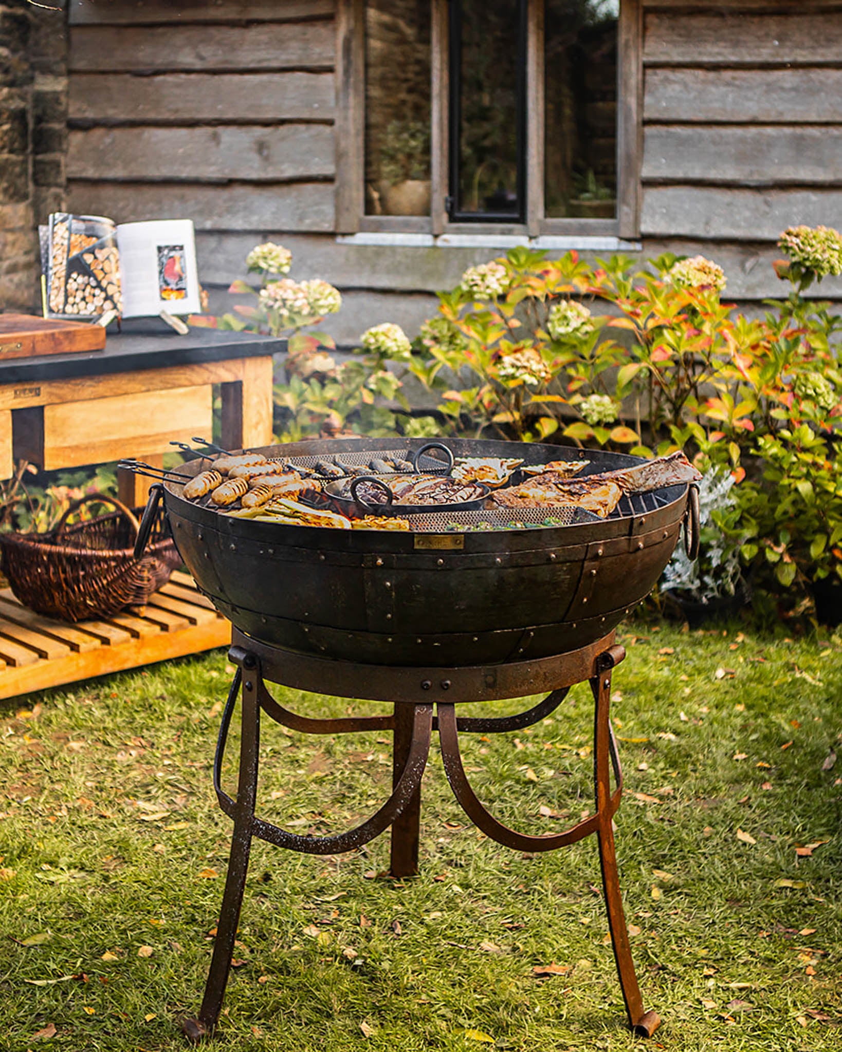 Medium sized BBQ fire pit with Kadai grill cooking sausages and meat in a garden beside a wooden house