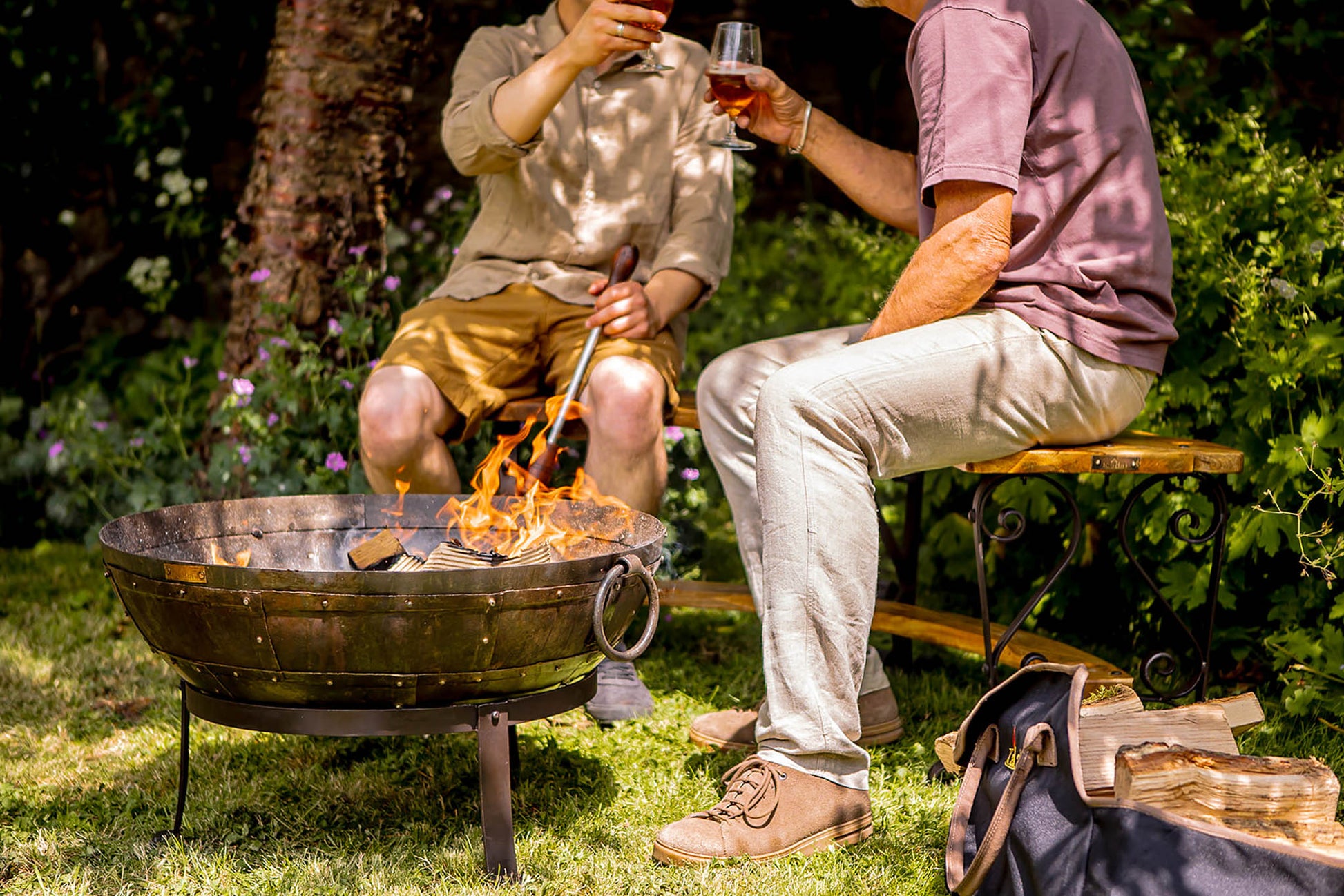 Medium sized charcoal BBQ fire bowl with flames as two people sit nearby enjoying drinks in garden