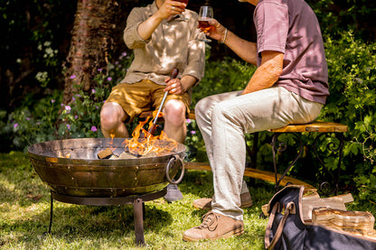 Medium sized charcoal BBQ fire bowl with flames as two people sit nearby enjoying drinks in garden