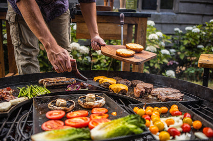 Person grilling vegetables and meats on a kadai barbecue grill outdoors.
