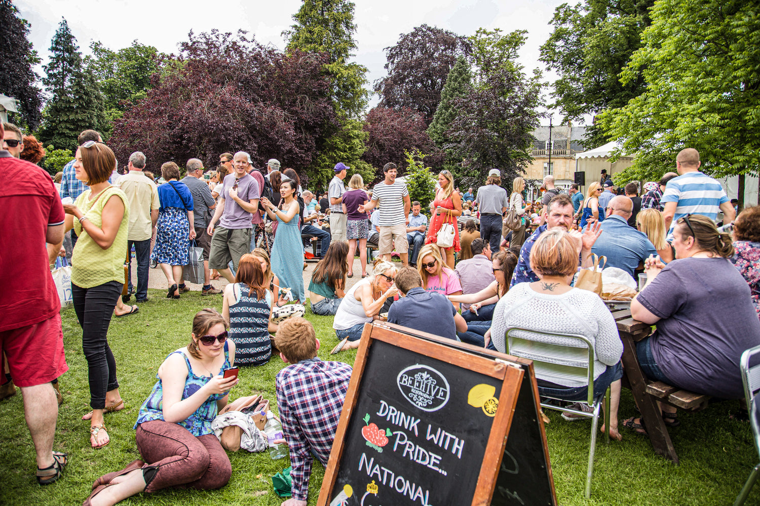 Outdoor event with people socializing in a park, featuring a chalkboard sign.
