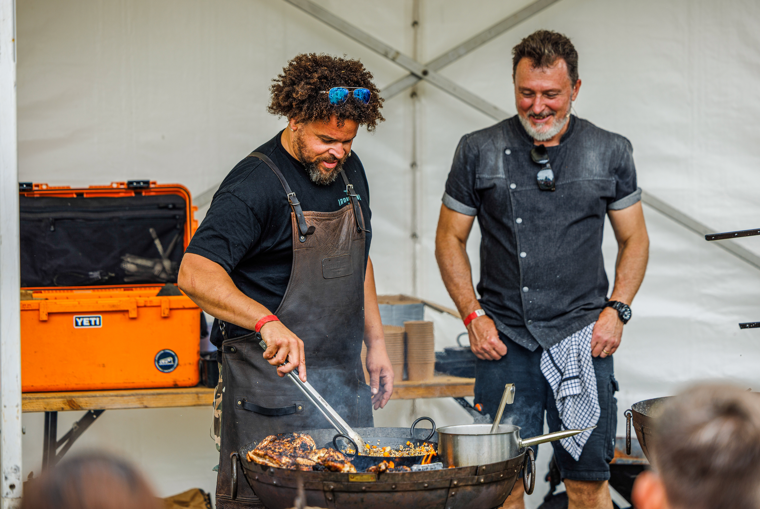 Two men cooking outdoors, one using tongs on a grill, with an orange cooler in the background.