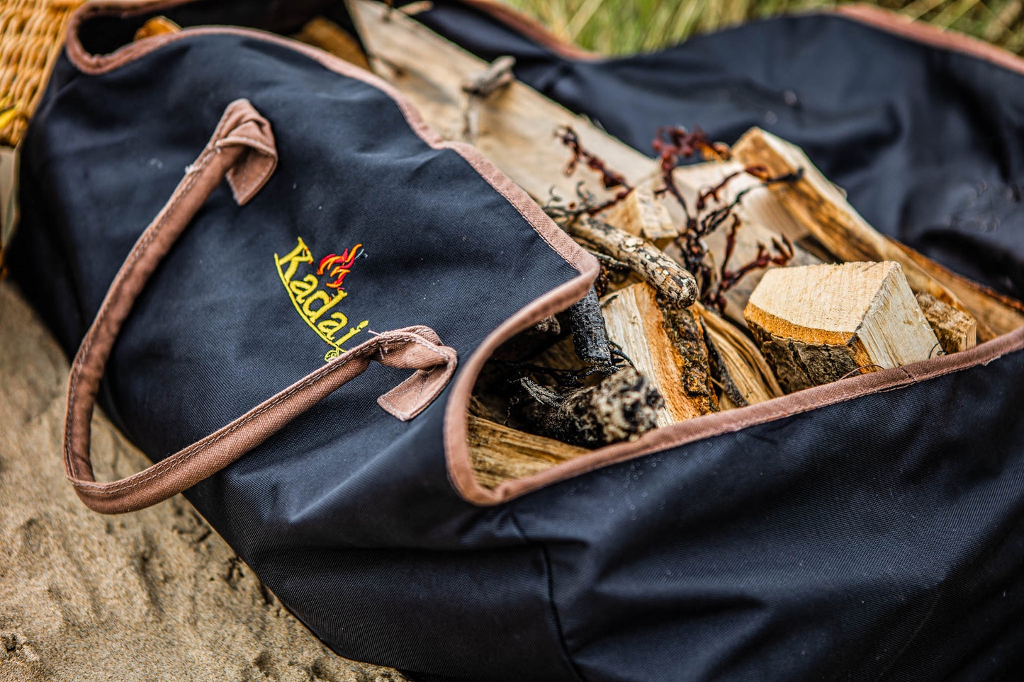 Black canvas log carrier filled with firewood and kindling, displayed outdoors on sandy ground.