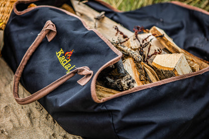 Black canvas log carrier filled with firewood and kindling, displayed outdoors on sandy ground.