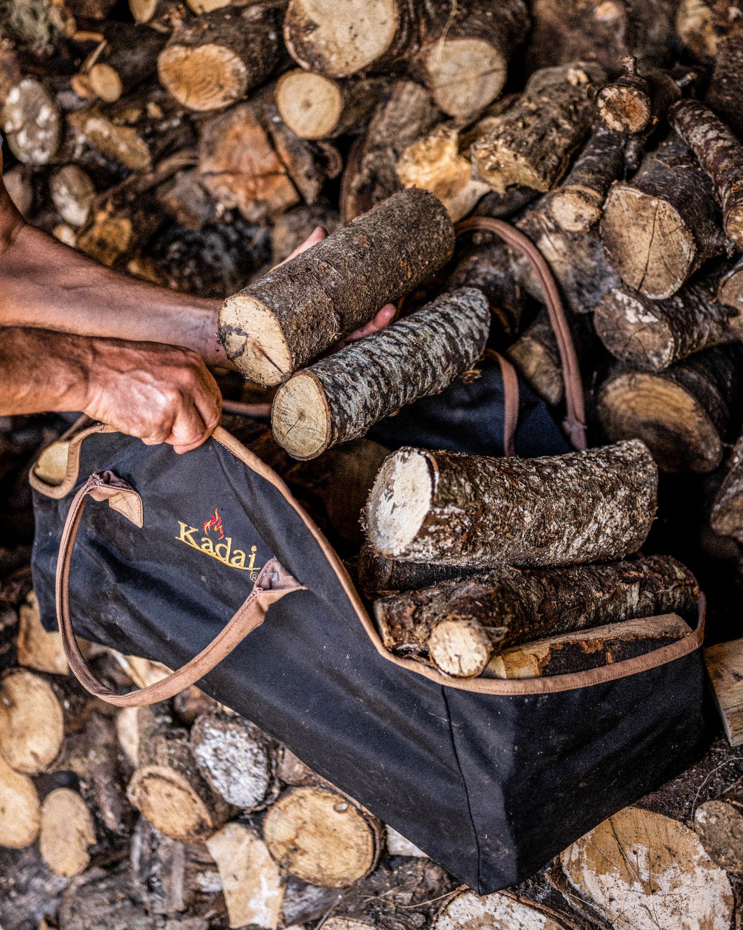 Person loading firewood logs into a black log carrier bag beside a stacked wood pile.