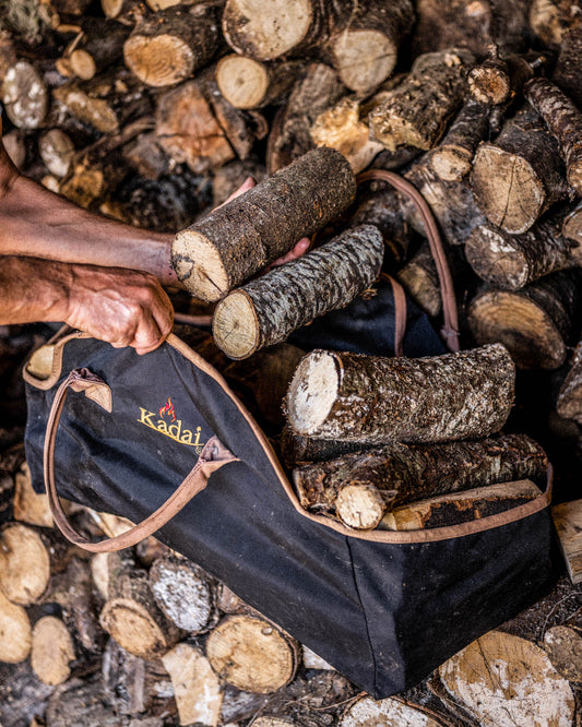 Person loading firewood logs into a black log carrier bag beside a stacked wood pile.