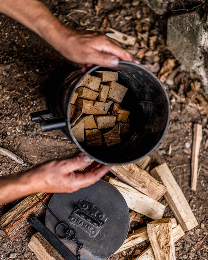Overhead view of hands filling a black steel charcoal maker with small wood pieces beside its lid, surrounded by split logs and kindling on the ground.