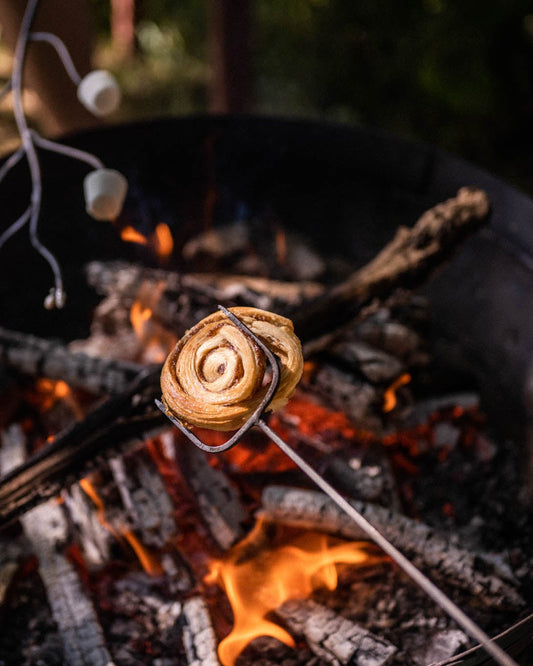 Pastry swirl toasted over glowing embers on a long metal toasting fork above an outdoor fire pit.
