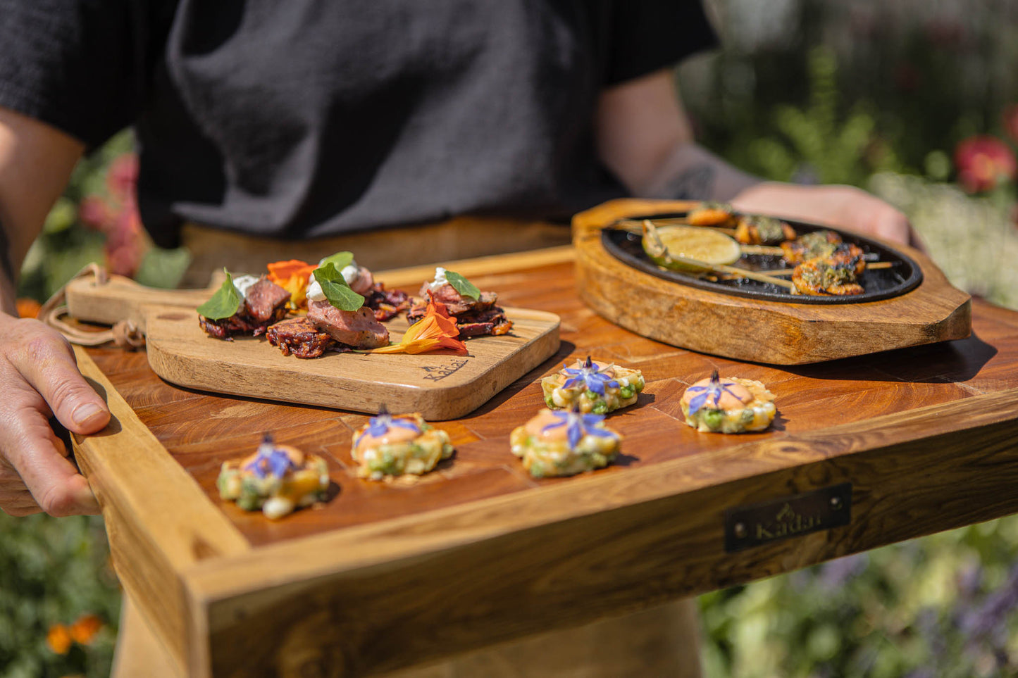 Person carrying a large wooden butcher’s block tray topped with elegant canapés, grilled bites, and colorful edible flower garnishes outdoors.