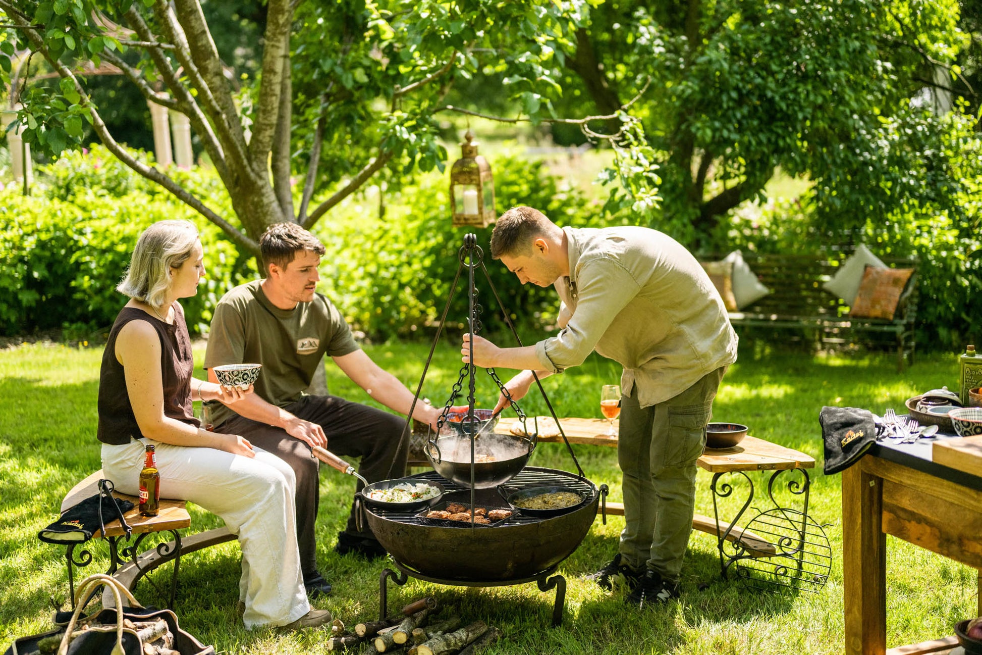 Friends gathered around a fire pit preparing food in a suspended cooking bowl with grill dishes outdoors