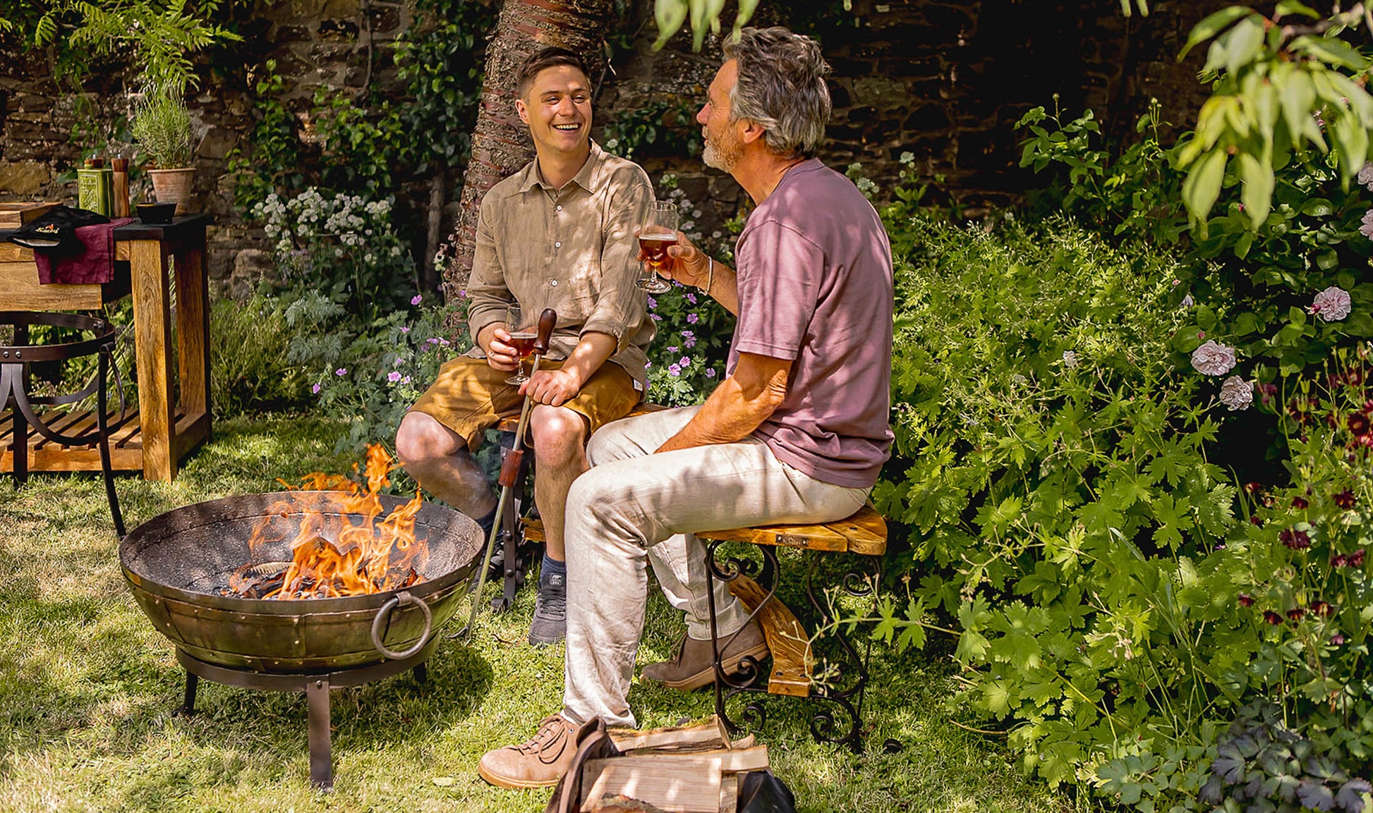 Kadai medium BBQ fire pit with burning logs, two men relaxing beside the fire with drinks in a garden setting