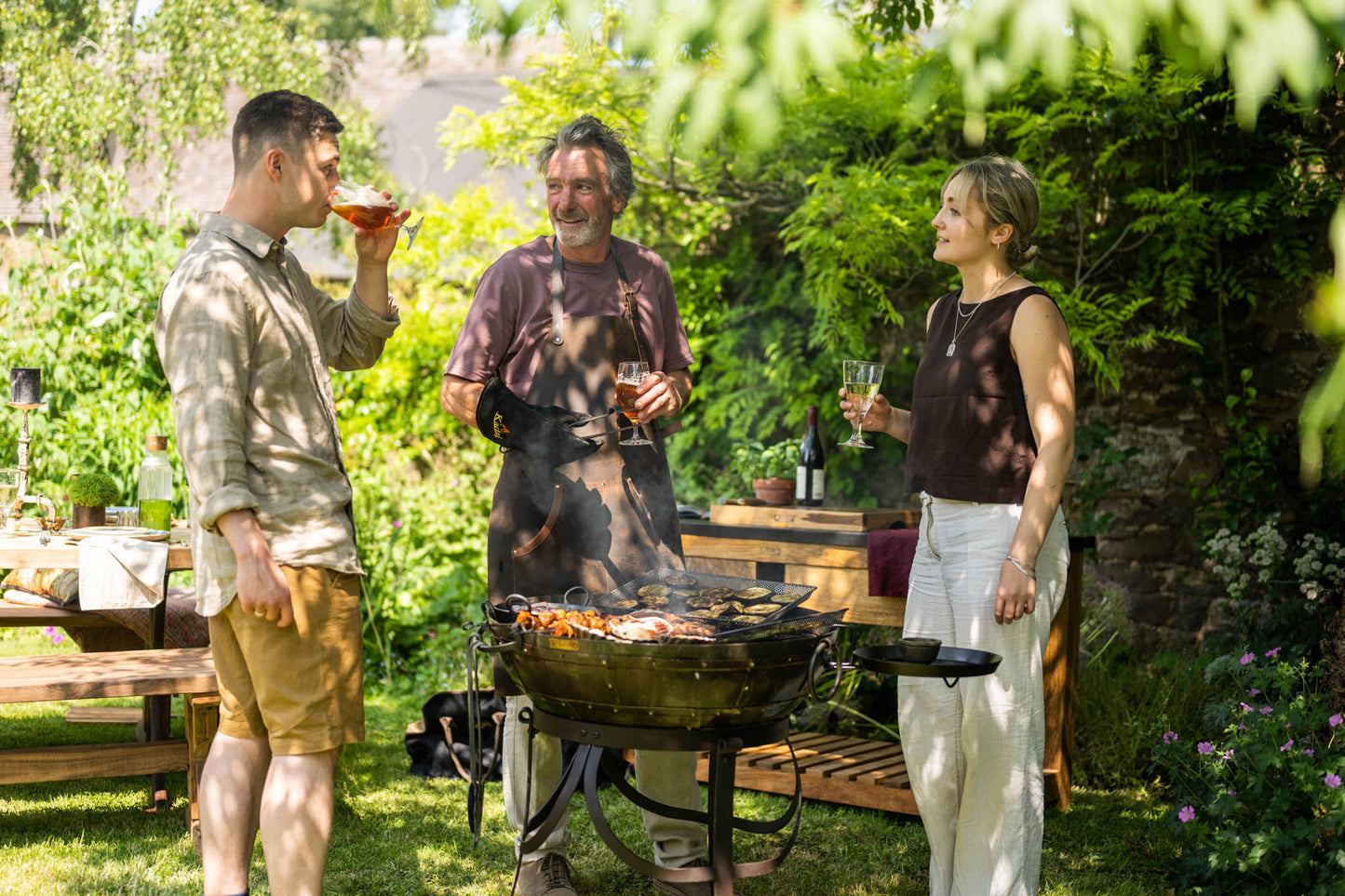 Medium BBQ fire pit with food cooking on a Kadai grill, three people standing around enjoying drinks in a garden