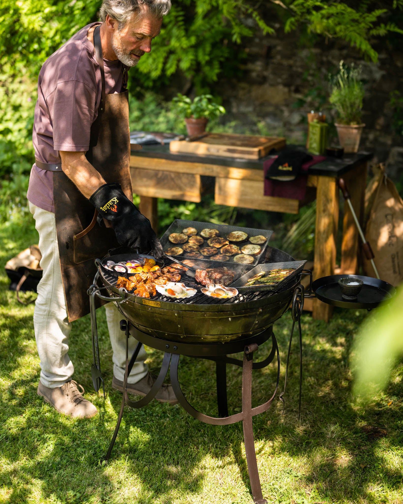 Kadai medium sized BBQ fire pit with food cooking on the grill, man tending the barbecue outdoors in a garden